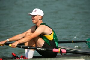 Male single scull rower wearing a green and yellow unisuit with white cap.