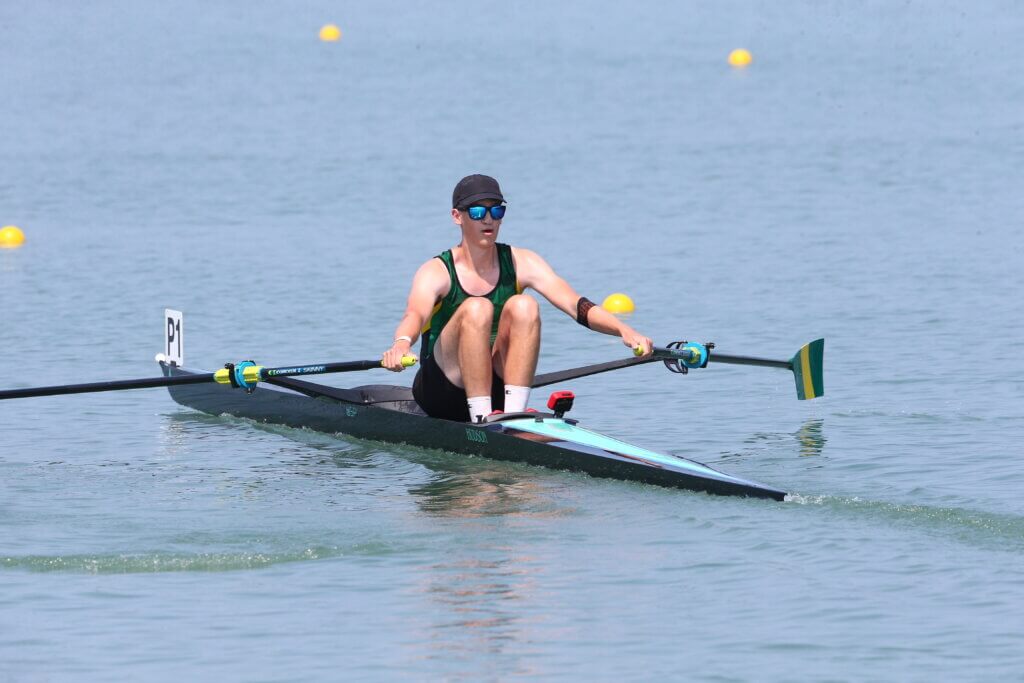 Rowing athlete with sunglasses and hat in open water rowing a dark blue boat sitting forward with oars in hand, ready to take a stroke