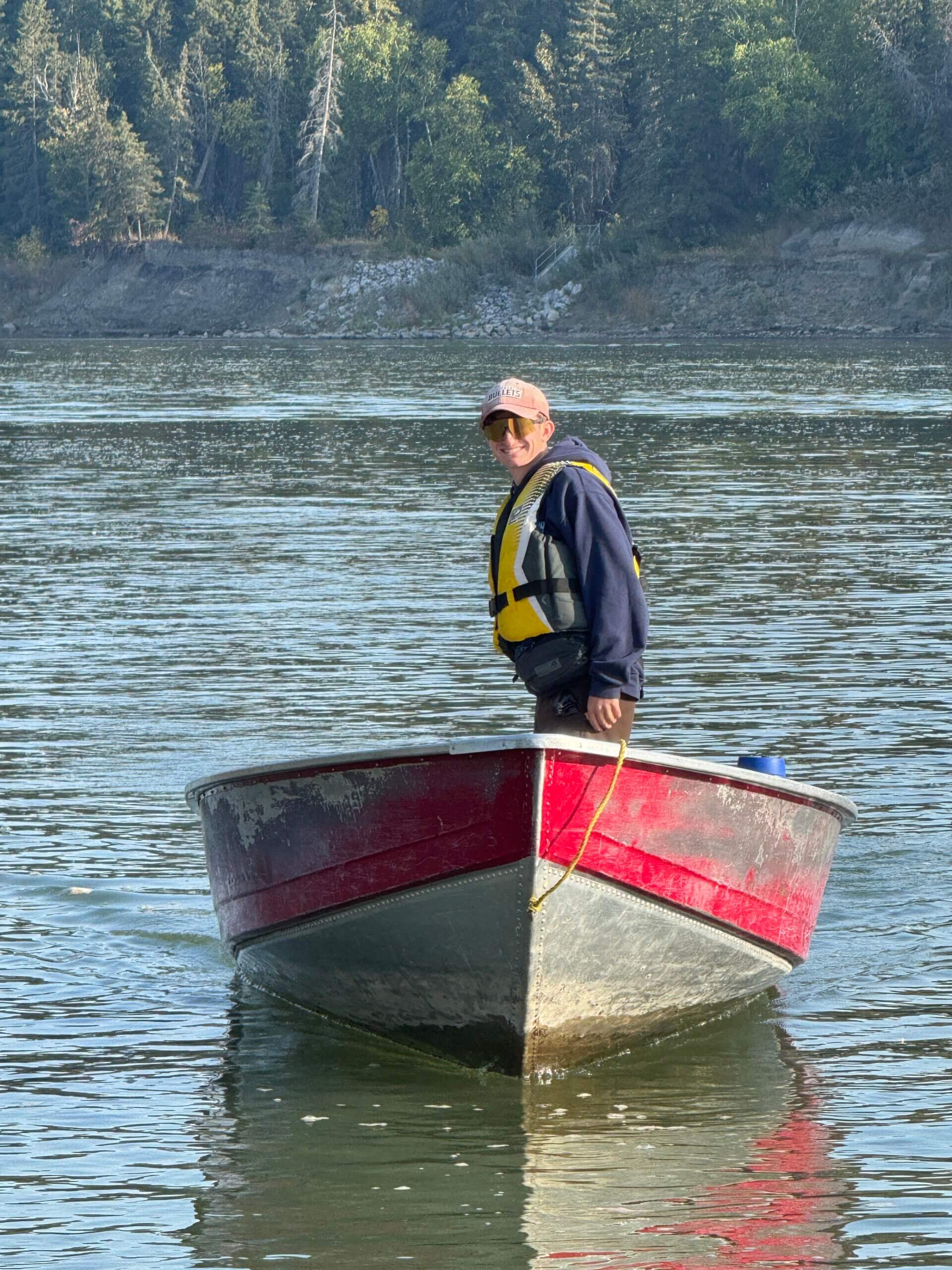 Rowing Coach standing in aluminum red trimmed motor boat on the North Saskatchewan River. Wearing a yellow lifejacket.